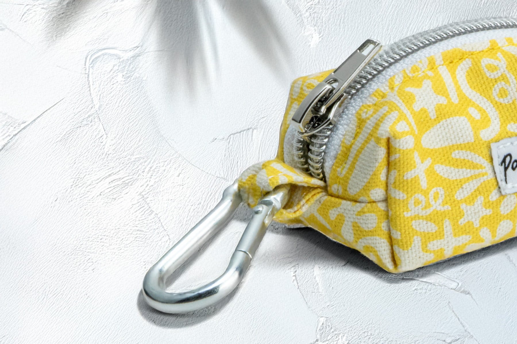 Gros plan sur le mousqueton et le fermeture éclaire de la pochette sacs à crottes Seaside pour chien. La pochette présente un motif marin abstrait blanc sur un fond jaune solaire. 