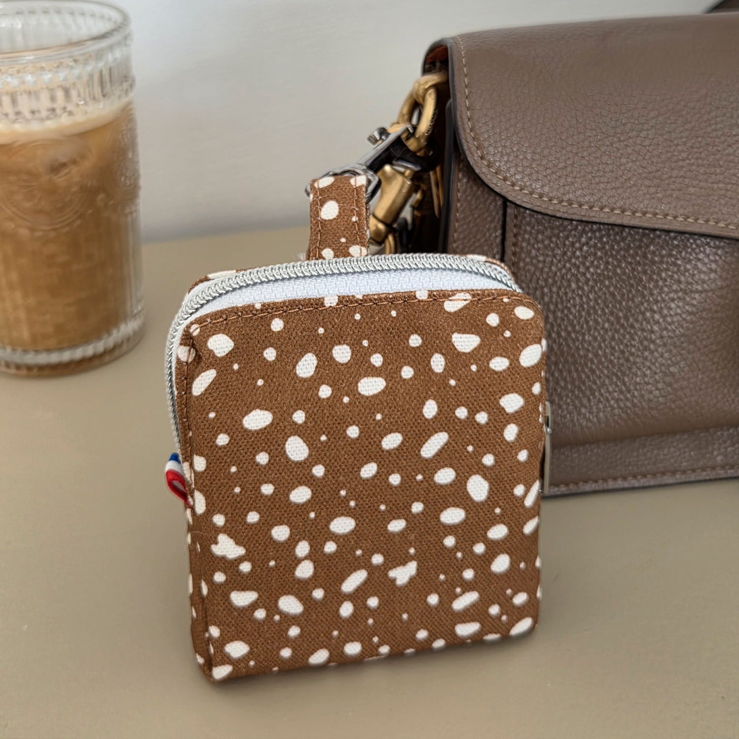Détail de la mini sacoche Freckles pour chien dans une ambiance lifestyle accroché à un sac à main. La mini sacoche présente un motif tacheté blanc sur fond marron.