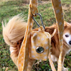 Gros plan sur la pochette sacs à crottes Peachy Daisies et son motif fleurs blanches et jaunes sur fond couleur pêche. La pochette est accroché à la laisse Peachy Daisies et l'on peut voir un petit chien en arrière plan